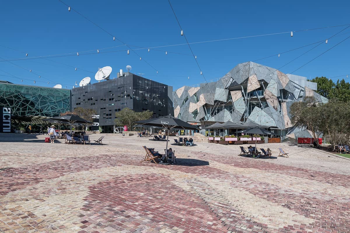 Der Federation Square in Melbourne an einem sonnigen Tag mit seinen markanten geometrischen Gebäuden und dem offenen Platz, auf dem Menschen unter Sonnenschirmen sitzen.