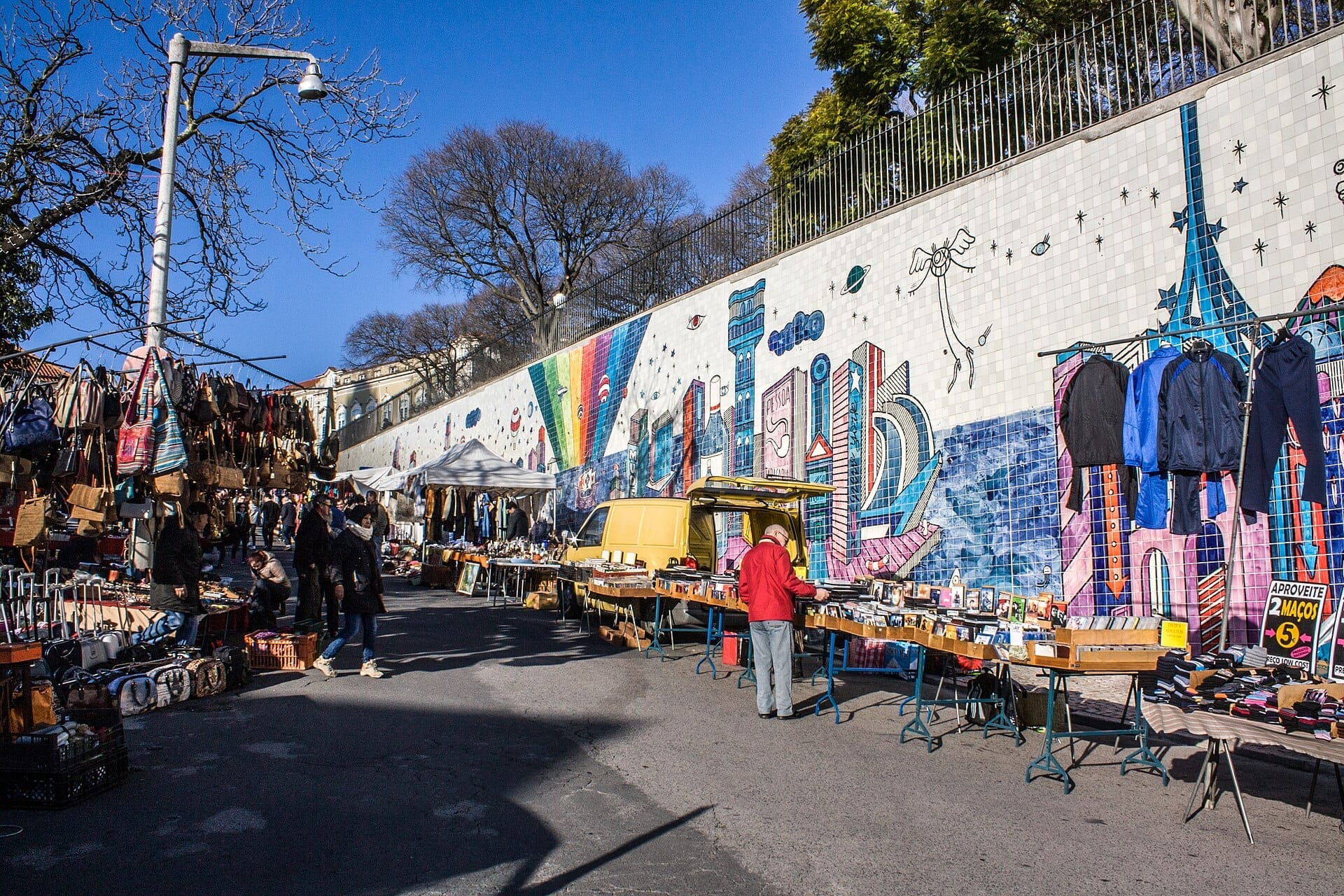 Bancarelle con articoli vintage e abbigliamento costeggiano la strada al mercato delle pulci Feira da Ladra, di fronte a un colorato murale nel quartiere Alfama di Lisbona.