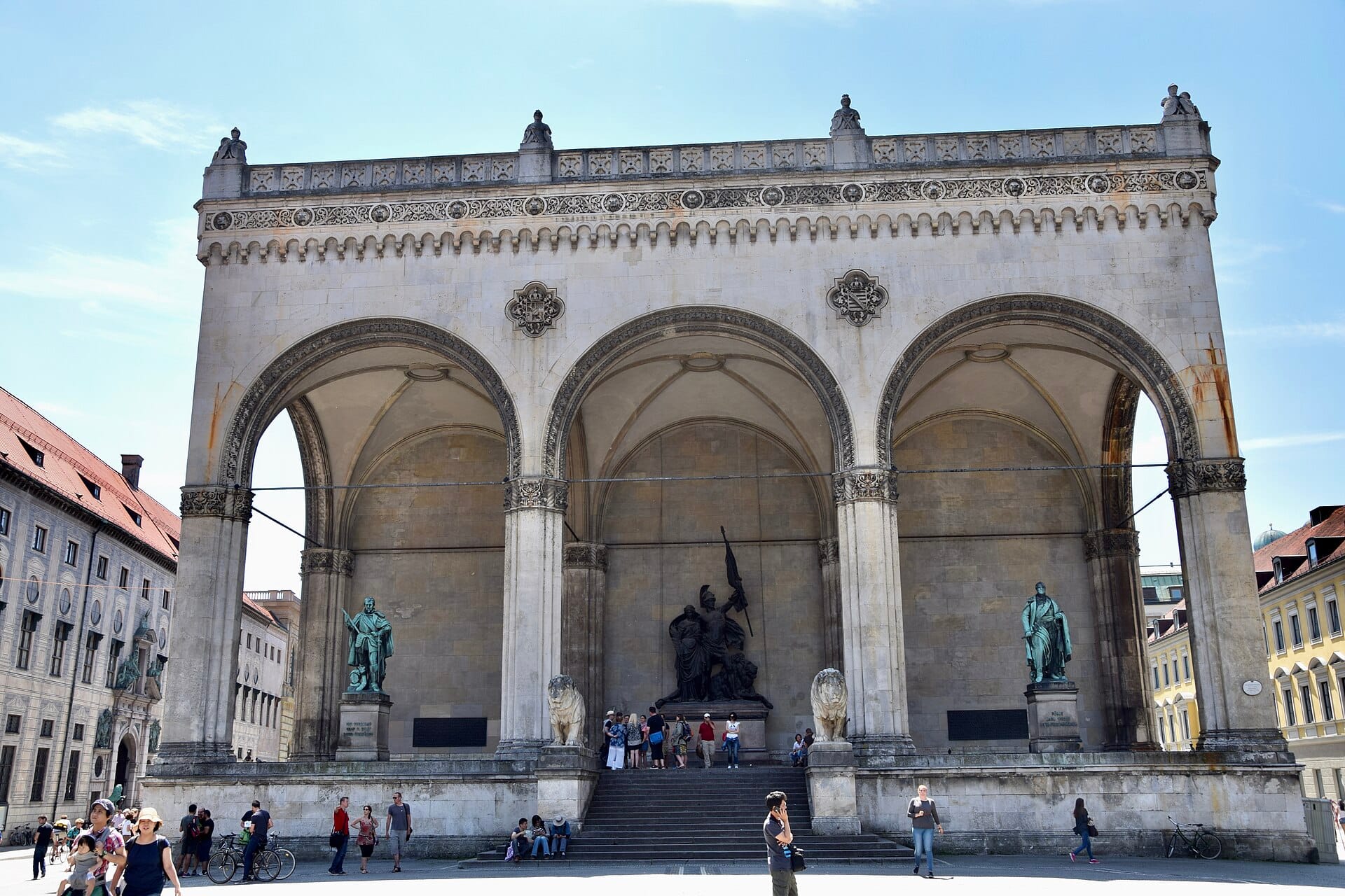 Feldherrnhalle ในมิวนิก มองจากด้านหน้าในวันที่แดดจ้า เห็นซุ้มโค้งขนาดใหญ่ 3 ช่อง รูปปั้น และผู้คนกำลังเดินอยู่ในจัตุรัส