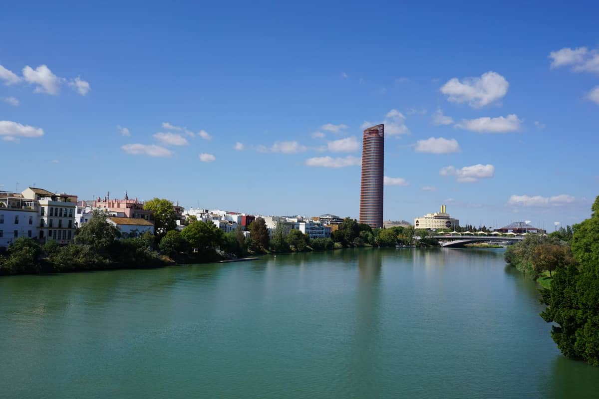วิวกว้างของริมแม่น้ำเมืองเซบียาพร้อมตึกระฟ้า Torre Sevilla อาคารในเมือง และสะพานข้ามแม่น้ำ Guadalquivir ใต้ท้องฟ้าสีฟ้า