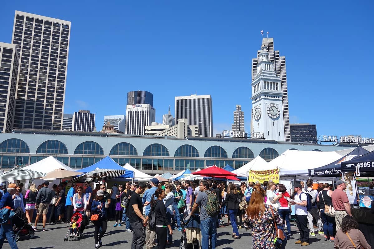 Folla che gode del Ferry Plaza Farmers Market di fronte all'iconico Ferry Building di San Francisco, con tende colorate e grattacieli della città sotto un cielo blu limpido.