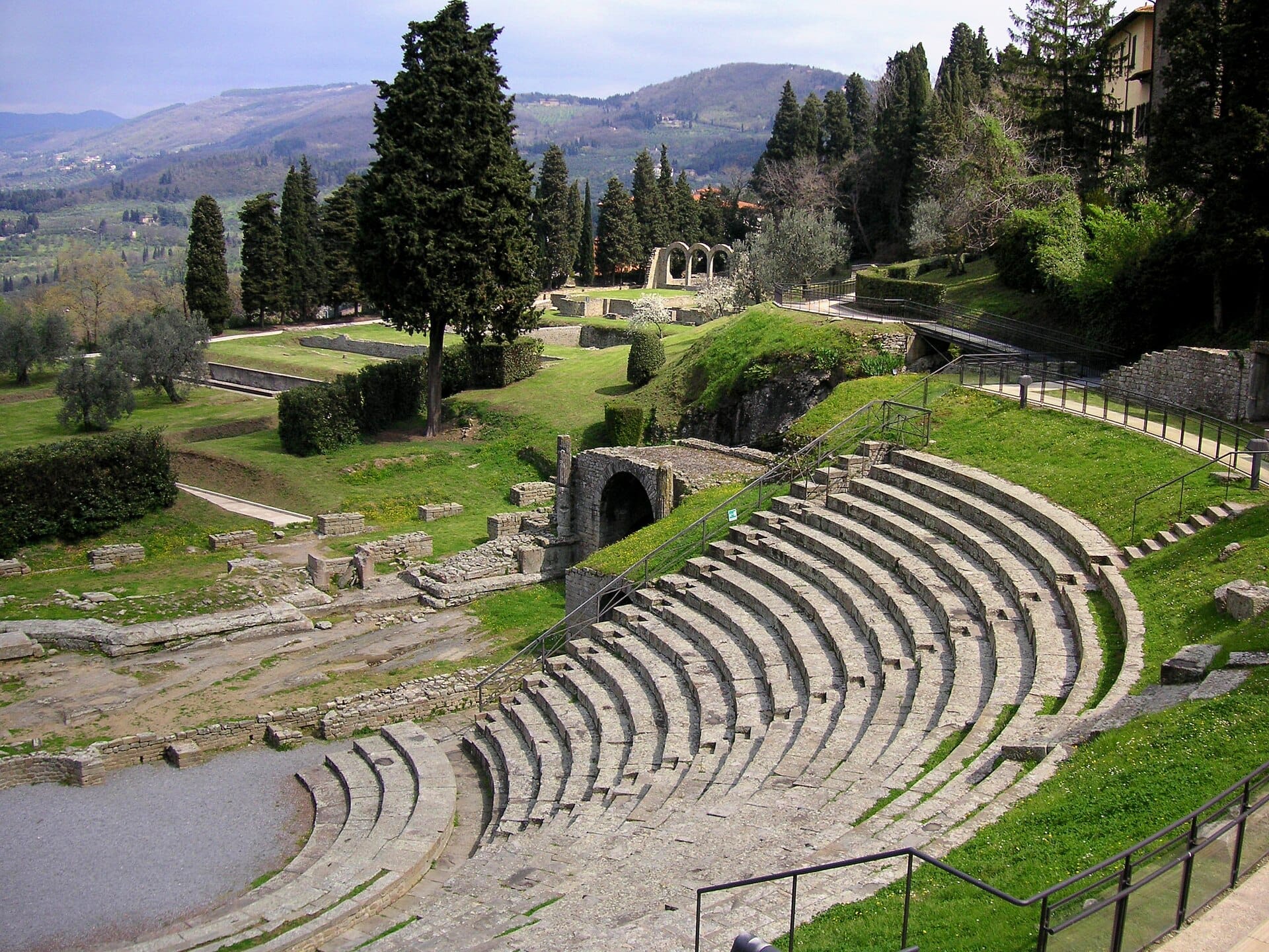 ที่นั่งหินของ Teatro Romano di Fiesole โค้งรอบเนินเขาสีเขียว พร้อมซากโบราณสถานและทิวทัศน์แคว้น Tuscany เป็นฉากหลัง