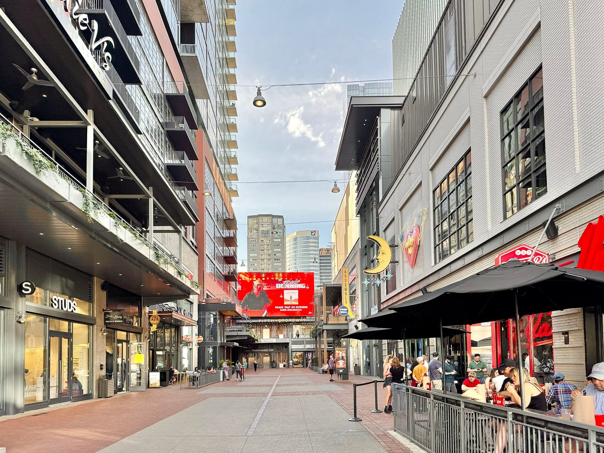 Vista a nivel de calle de Fifth + Broadway en Nashville con edificios modernos, escaparates de vidrio, comedor al aire libre y una valla digital bajo un cielo azul.