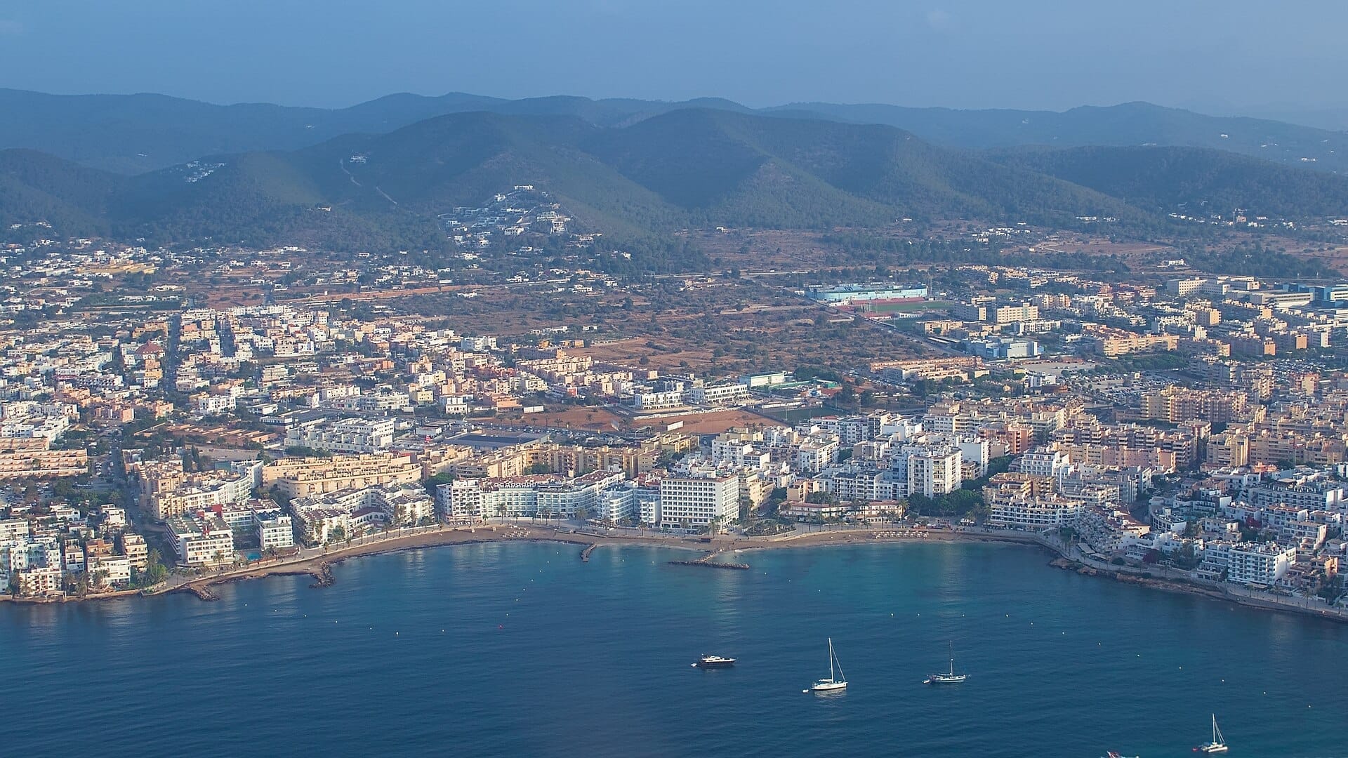 Vista aerea della spiaggia di Figueretas a Ibiza Town, con edifici urbani, riva sabbiosa, acque blu calme, barche e colline verdi sullo sfondo.