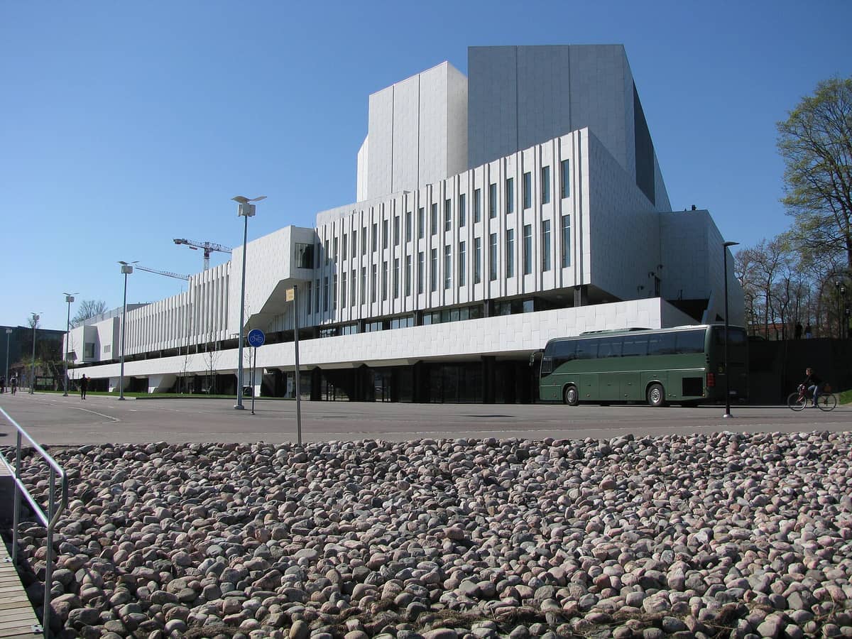 A marcante fachada de mármore branco e o design modernista geométrico do Finlandia Hall se destacam sob um céu azul claro, com um ônibus turístico estacionado nas proximidades.