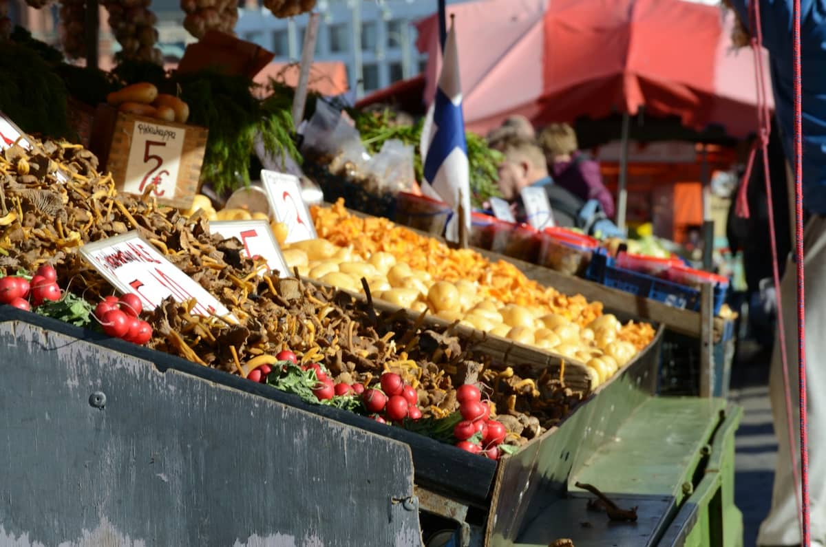 Barraca de mercado ao ar livre em Helsinki com cogumelos silvestres, frutas vermelhas e produtos locais sob toldos vermelhos e uma bandeira finlandesa visível.
