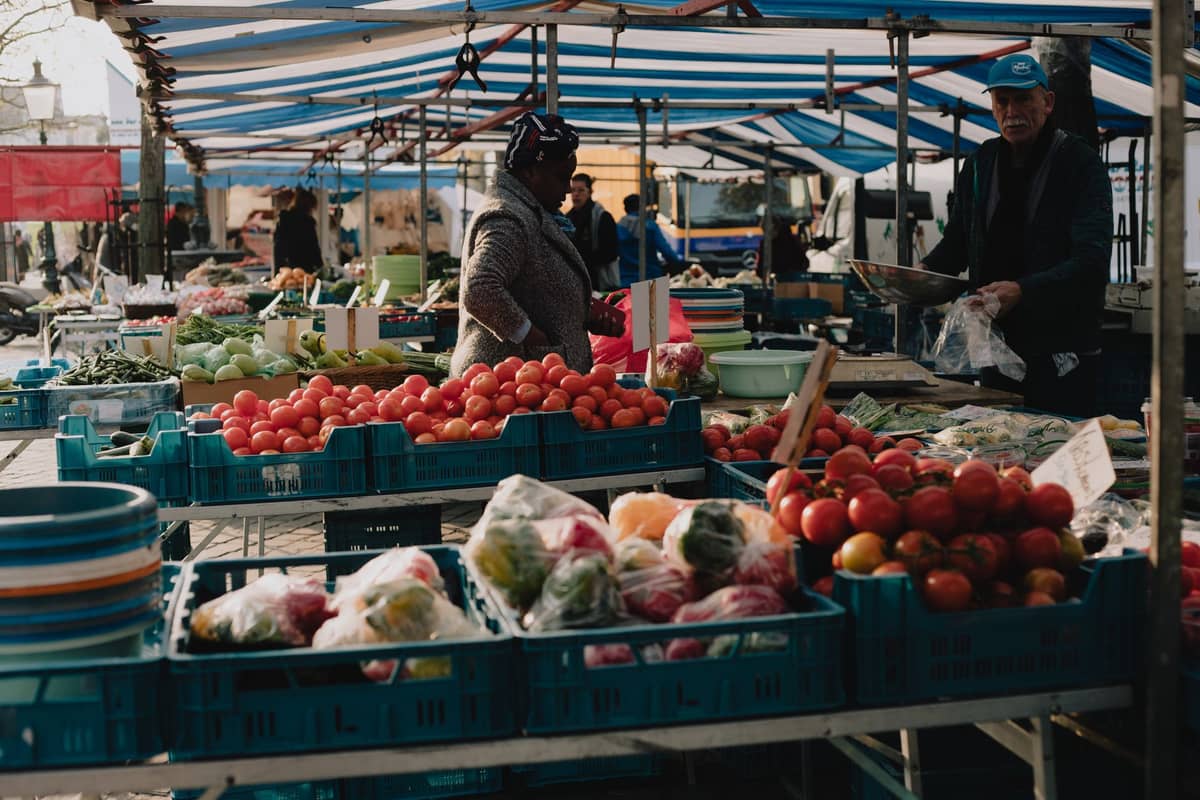 Cena de mercado ao ar livre em clima mais frio com barracas exibindo produtos locais frescos, tomates, pimentões e verduras, sob uma tenda listrada em uma cidade europeia.
