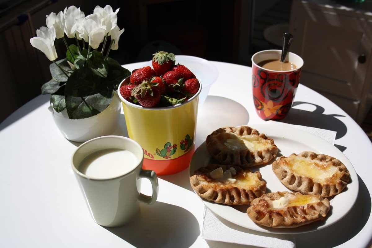 Mesa com comidas tradicionais finlandesas, incluindo tortas carelianas, morangos, café e leite à luz natural do dia.