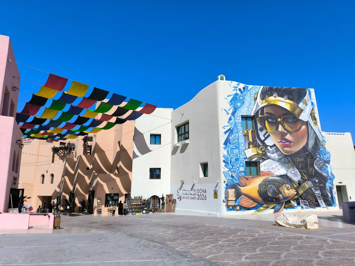Innenhof des Fire Station: Artist in Residence in Doha mit markanten pastellfarbenen Gebäuden, einem großen zeitgenössischen Wandbild und bunten Fahnen unter einem strahlend blauen Himmel.