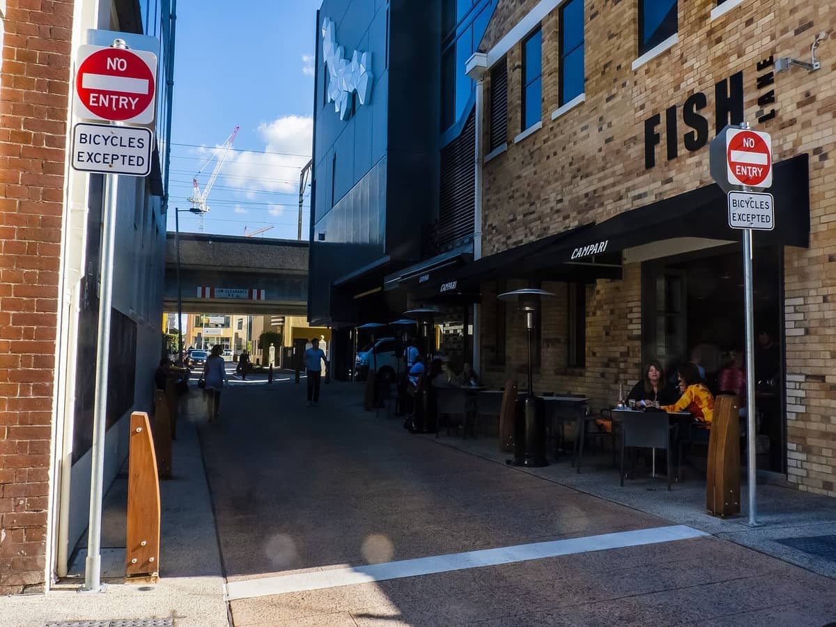 Outdoor diners sit at tables beside brick buildings under signage for Fish Lane, with pedestrians and street signs visible on a sunny afternoon.