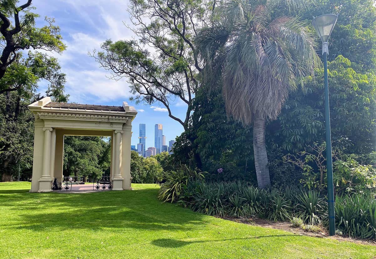 Un pabellón con columnas clásicas y techo ornamental se alza entre amplias zonas verdes y árboles en Fitzroy Gardens, con los rascacielos de Melbourne al fondo.