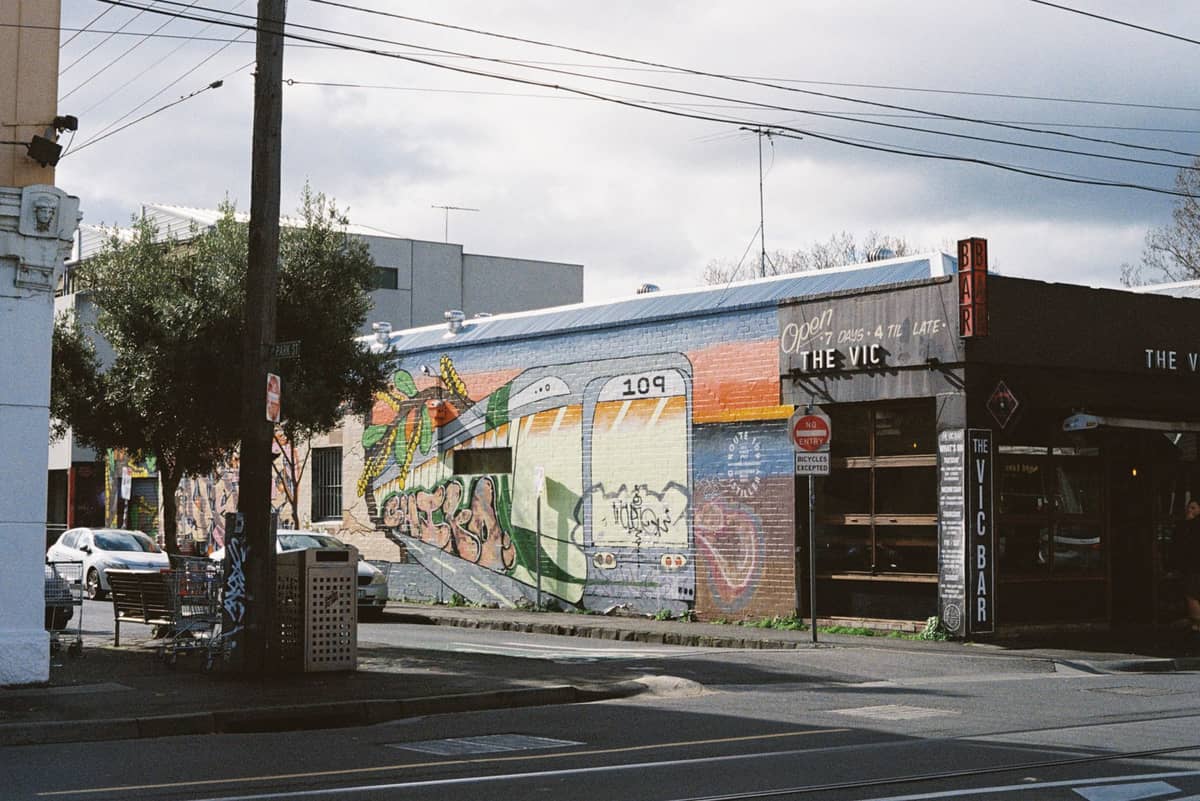 ถนนใน Fitzroy หรือ Collingwood กับภาพกราฟฟิตี้รถรางสีสันสดใสข้างอาคาร ใกล้บาร์และต้นไม้