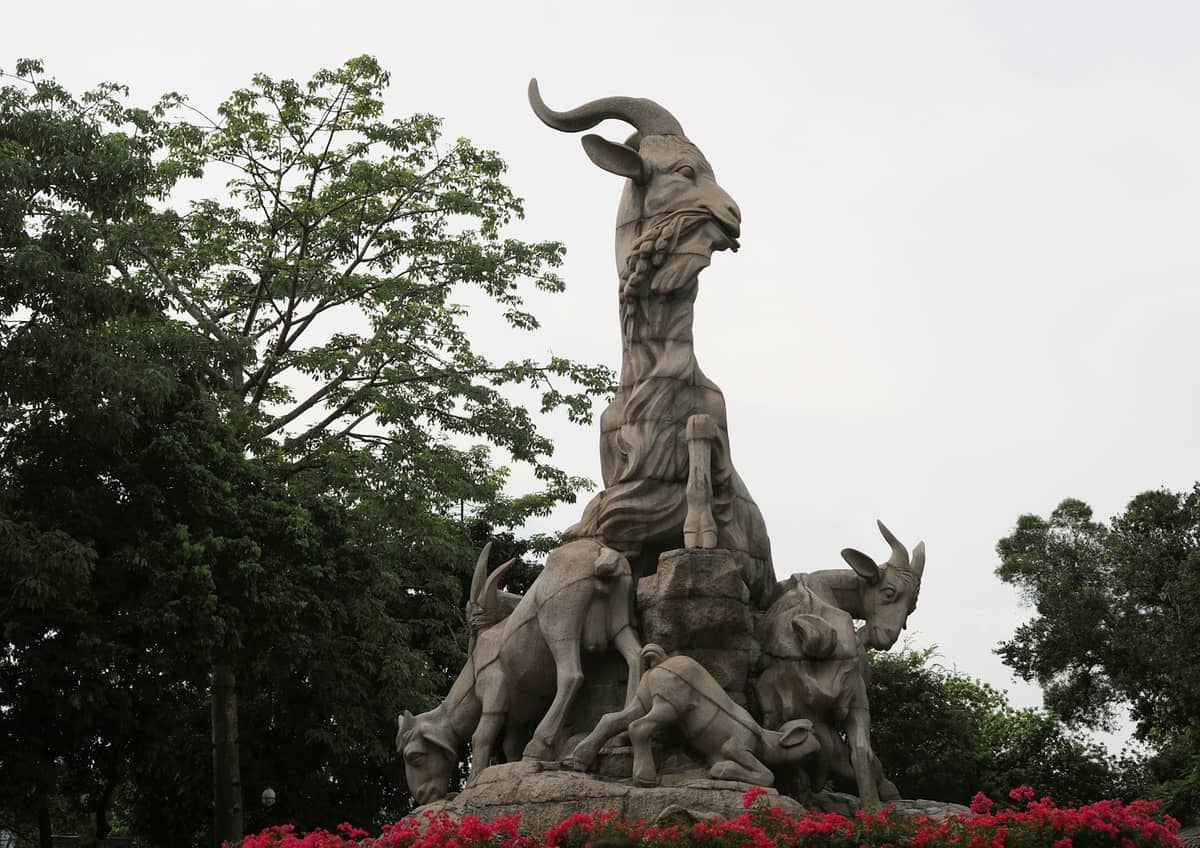 The Five Rams Statue in Yuexiu Park, Guangzhou, surrounded by lush green trees and vibrant red flowers under an overcast sky.