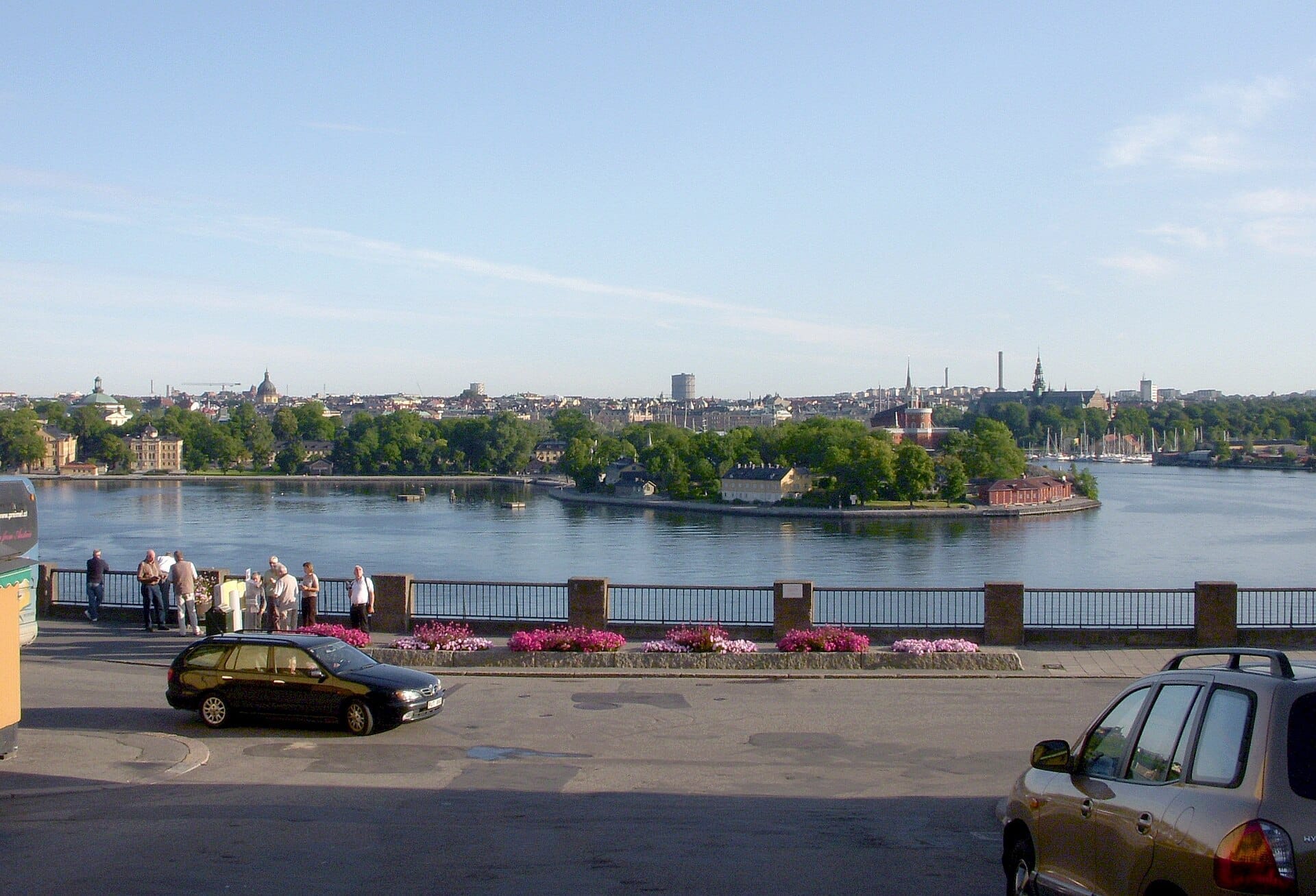 วิวจาก Fjällgatan ที่มีผู้คนชื่นชมเส้นขอบฟ้าของ Stockholm ข้ามน้ำ โดยมีดอกไม้สดใสและรถยนต์จอดอยู่ในบริเวณเบื้องหน้า