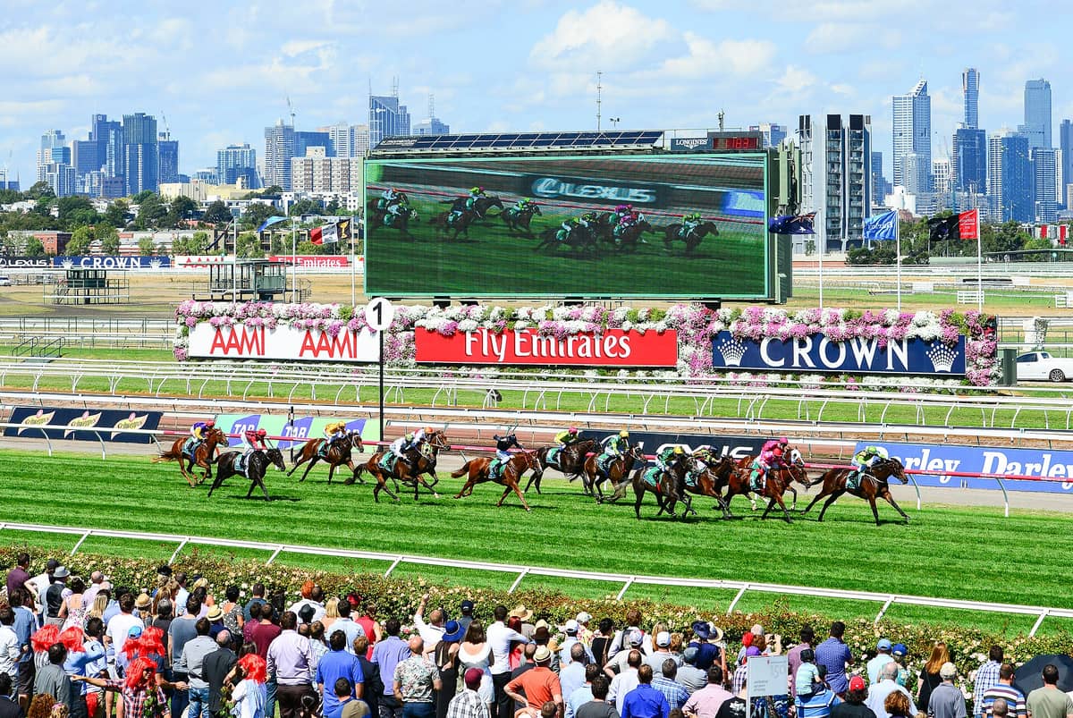 Vista panorámica de una carrera en el Hipódromo de Flemington, con caballos en la pista, una tribuna llena, flores y el horizonte de Melbourne al fondo.