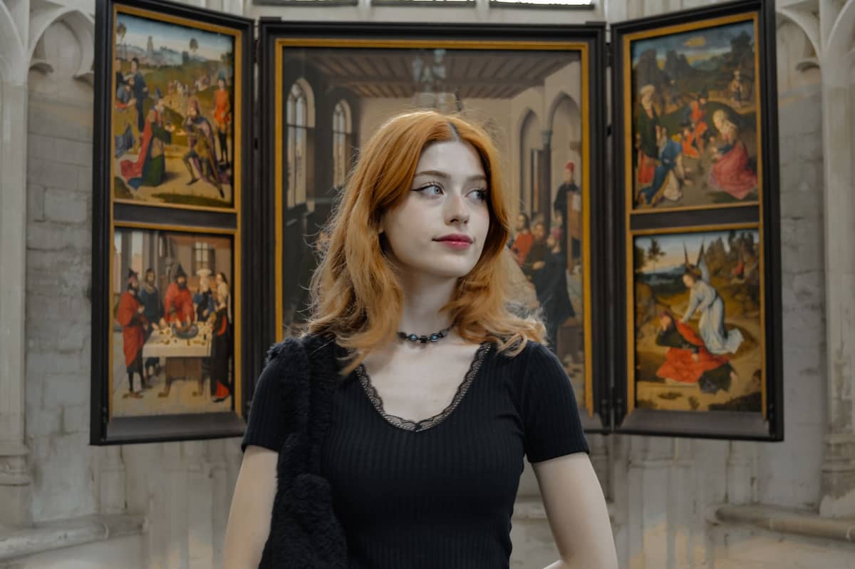 Young woman stands inside a church before a multi-paneled altarpiece featuring scenes in the style of Flemish Primitive art.