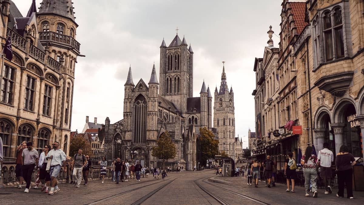 Street view of Ghent with St Nicholas' Church and the Belfry, lined with historic Flemish buildings and people walking.