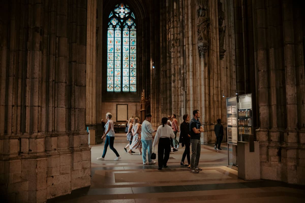 Visitors and tourists walking inside an ornate cathedral with tall stone columns and stained glass windows.