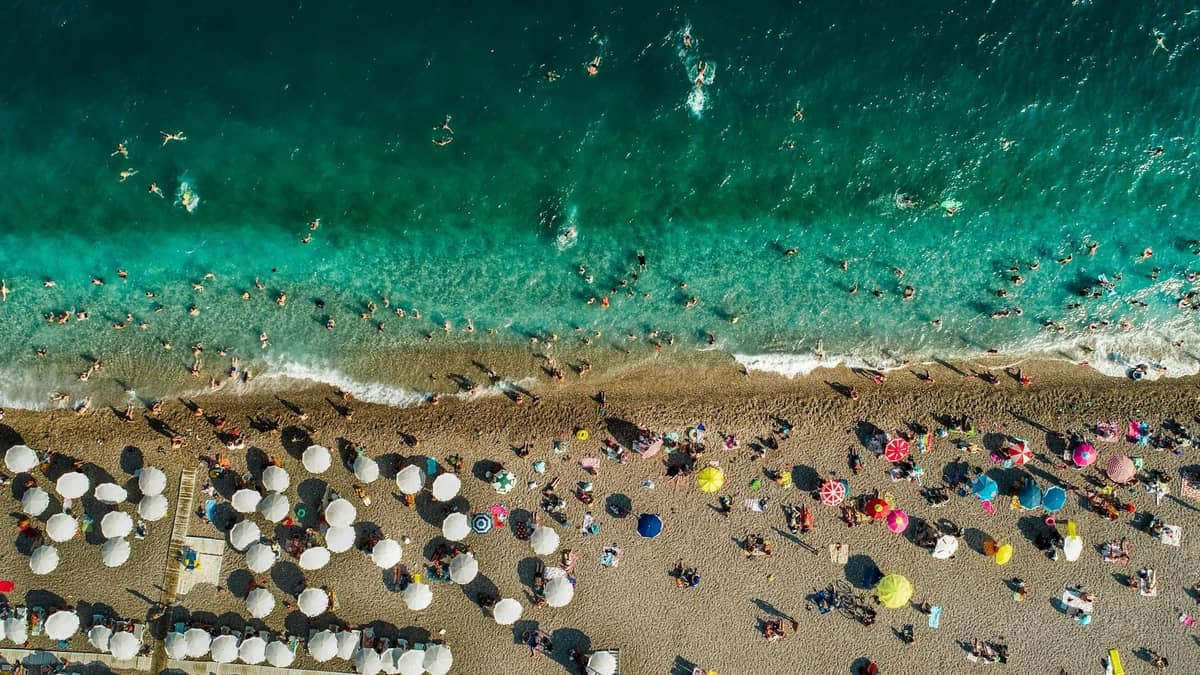 Aerial view of a crowded Antalya beach with sun umbrellas and swimmers, showing vibrant summer activity.