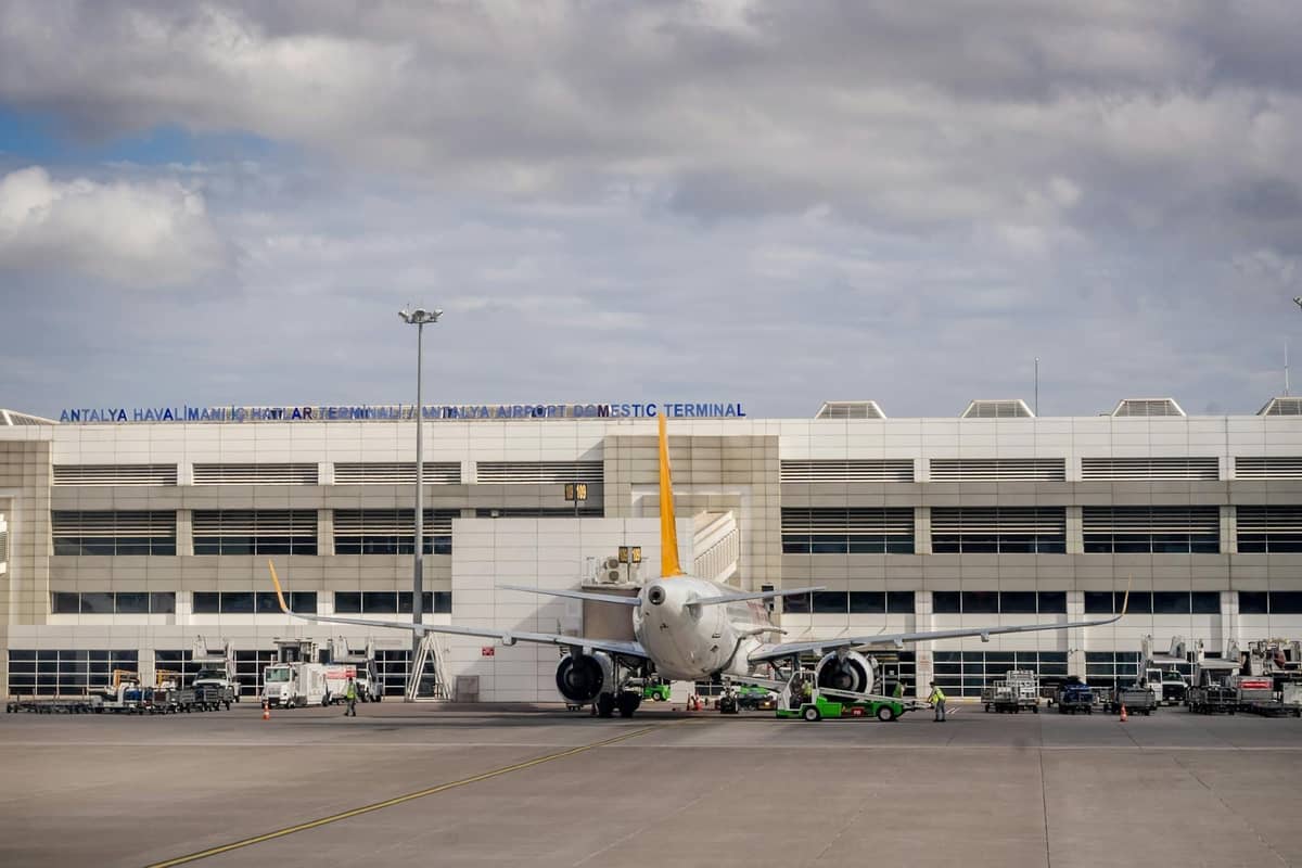 A commercial airplane at the terminal of Antalya Airport with the building facade and airport name visible.