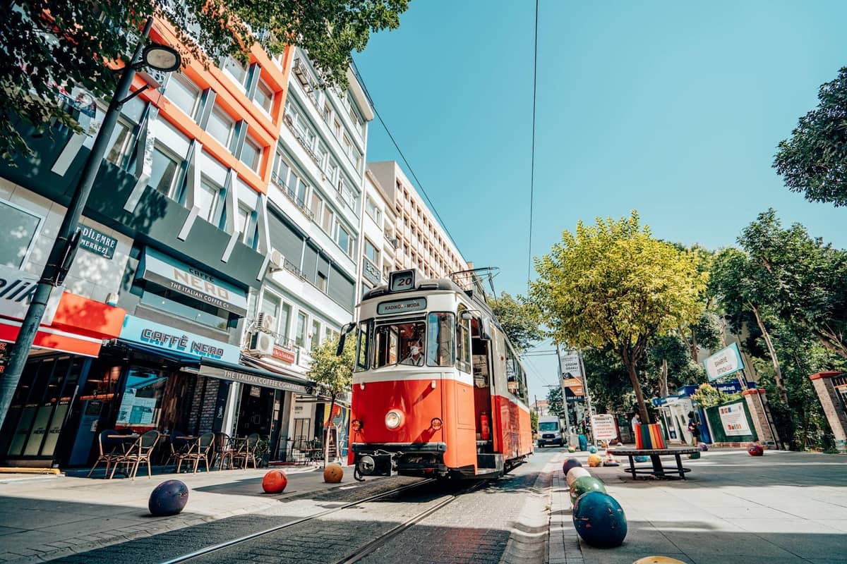 Daytime view of an iconic Antalya tram passing through a central city street with modern buildings, outdoor cafes, and trees lining the tracks.