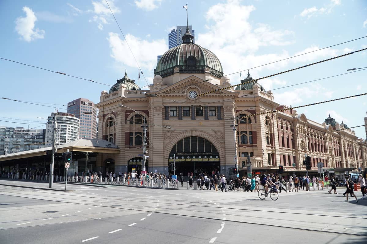Vista diurna y amplia de la icónica fachada con cúpula y entrada en arco de la Estación de Flinders Street, con personas cruzando la concurrida intersección y edificios de la ciudad al fondo.