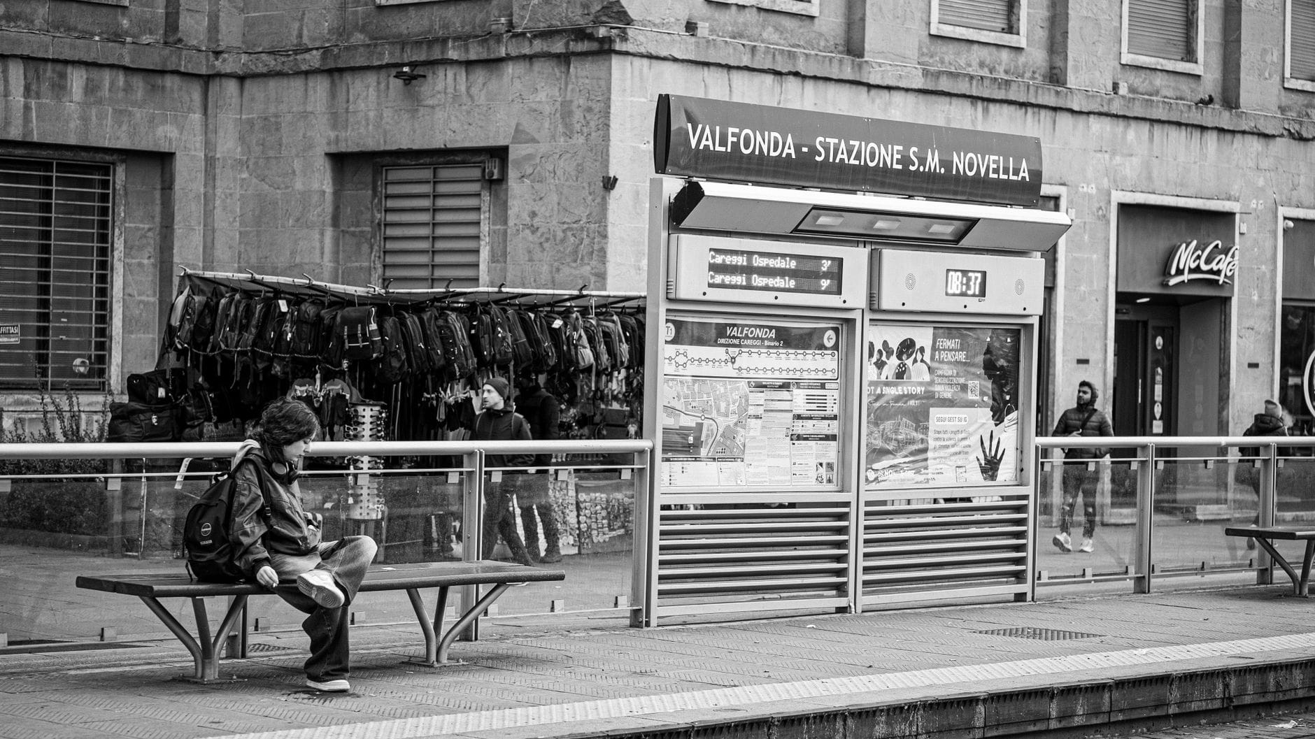 ภาพขาวดำของบริเวณทางเข้าด้านนอกของ Valfonda-Stazione S.M. Novella ในเมืองฟลอเรนซ์ โดยมีผู้คนและซุ้มข้อมูลให้เห็น