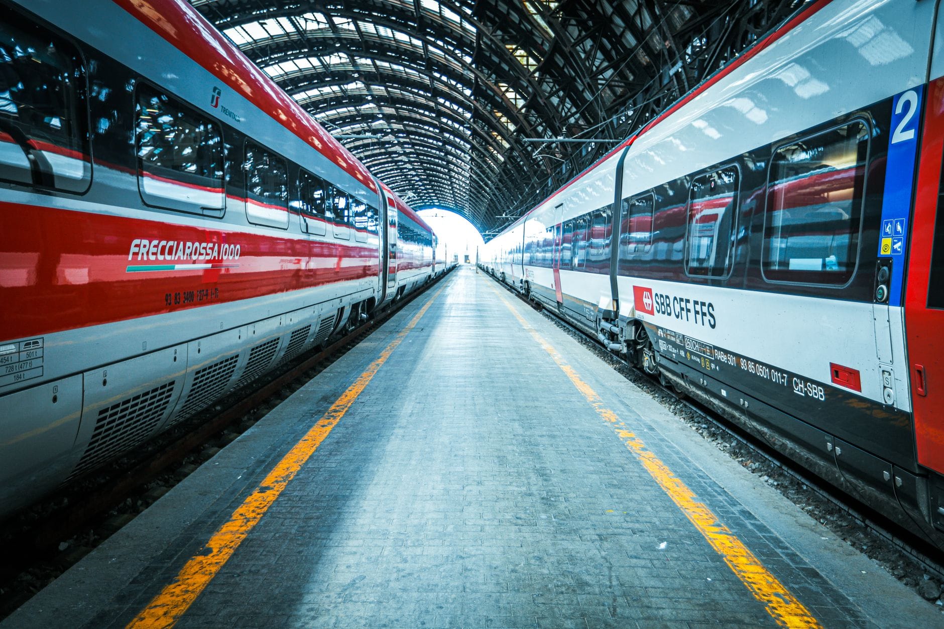 รถไฟความเร็วสูงสองขบวนที่สถานีในอิตาลี รวมถึง Frecciarossa สีแดง ภายใต้หลังคาโค้งขนาดใหญ่ หันหน้าไปยังชานชาลากว้างที่ว่างเปล่า