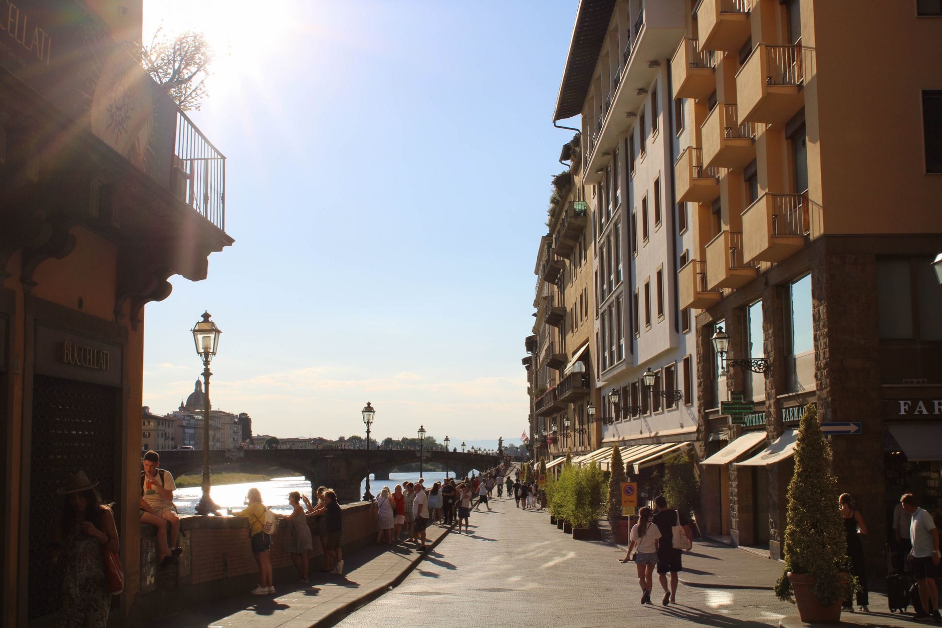 ผู้คนเดินไปตามถนนคนเดินกว้างริมแม่น้ำในเมือง Florence พร้อมอาคารประวัติศาสตร์และแสงแดดเป็นฉากหลัง