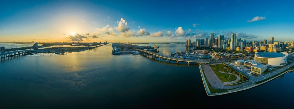 Panoramic aerial view of downtown Miami skyline at sunrise with causeways extending over Biscayne Bay toward the horizon.
