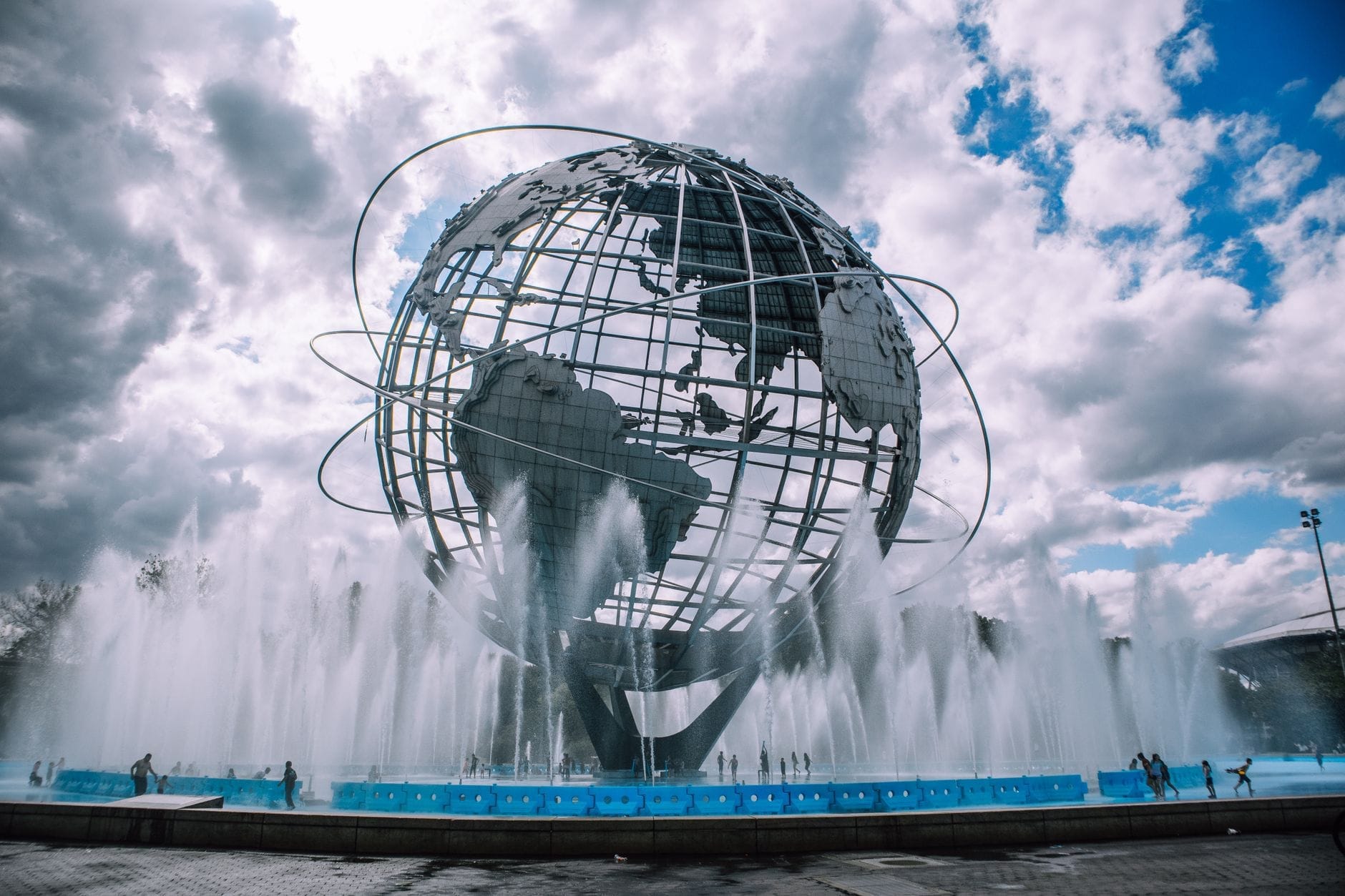 L'iconica Unisfera circondata dalle fontane al Flushing Meadows Corona Park sotto un cielo drammatico con qualche nuvola, mentre i visitatori passeggiano nei dintorni.