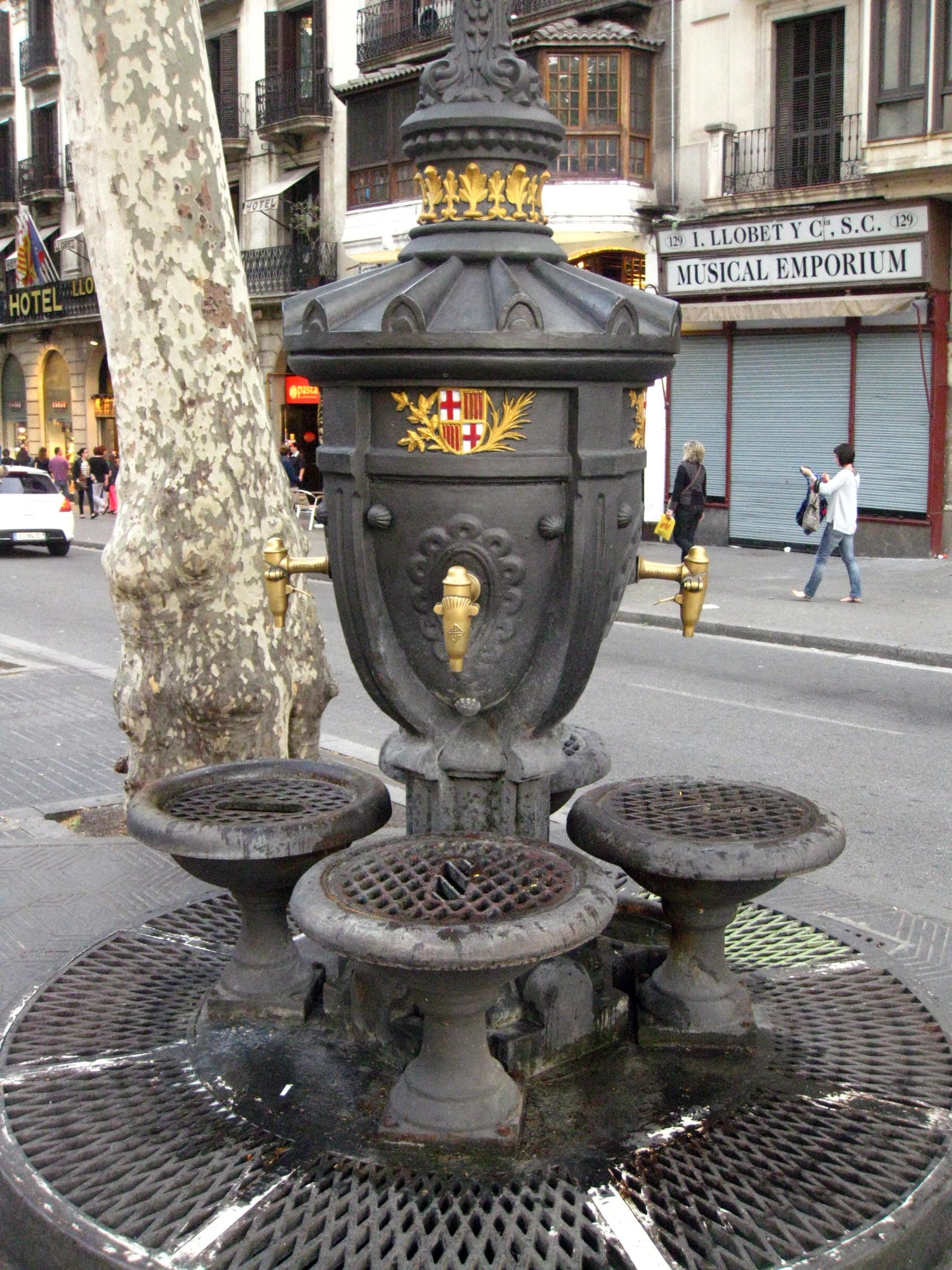 The Font de Canaletes, an ornate black cast-iron fountain with gold details, stands beside a tree on a bustling Barcelona street.