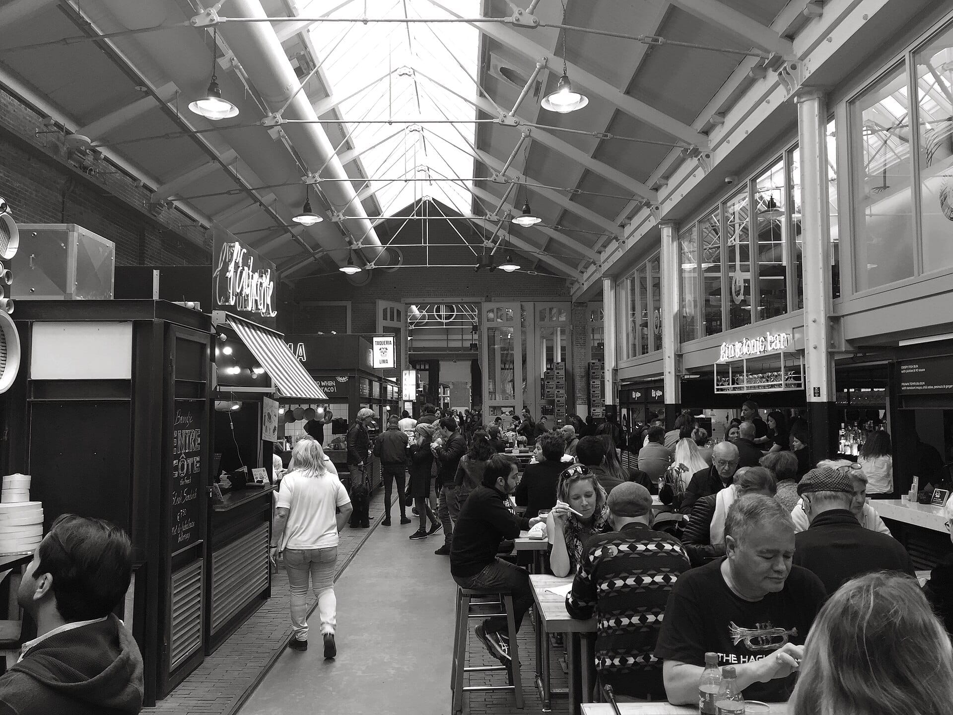 Vista interior de Foodhallen Amsterdam con techos altos, arquitectura industrial, puestos de comida y visitantes sentados en mesas comunitarias en un ambiente animado.