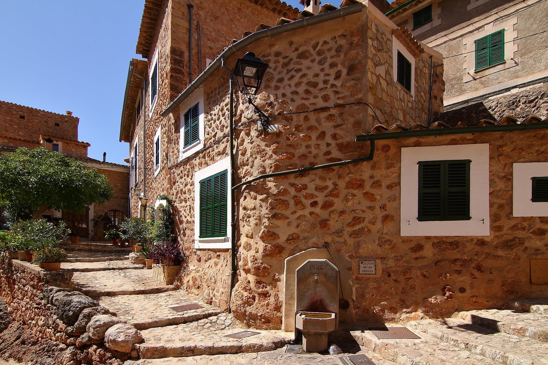 Case in pietra con persiane verdi costeggiano una strada acciottolata e soleggiata a Fornalutx, Maiorca, con piante in vaso e una vecchia fontana visibili.