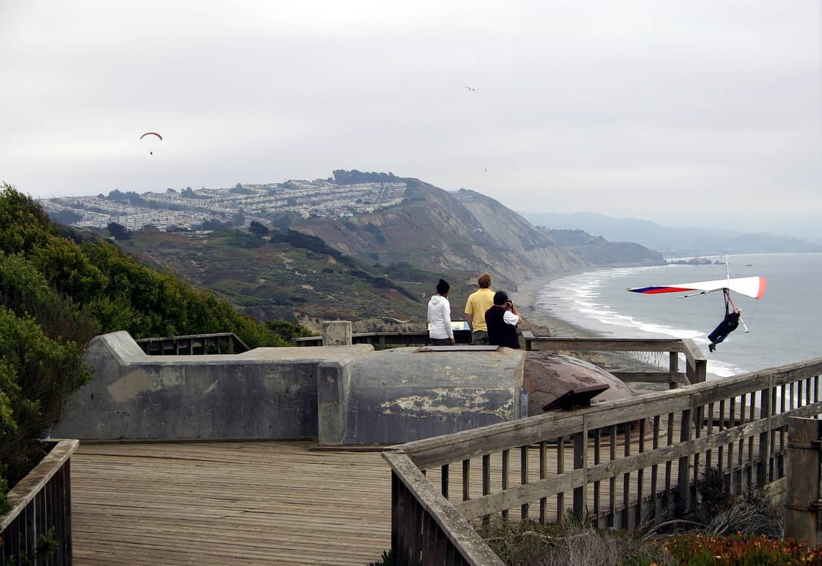 Blick auf die Küstenklippen von Fort Funston mit drei Personen, die den Ozean beobachten, einem Hängegleiter, einem Sandstrand und einem entfernten Stadtviertel von San Francisco.