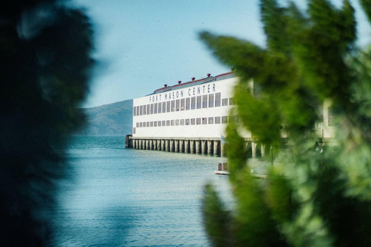 El edificio de Fort Mason Center frente al mar visto entre vegetación, con su nombre visible en la parte superior y la bahía de San Francisco en primer plano.