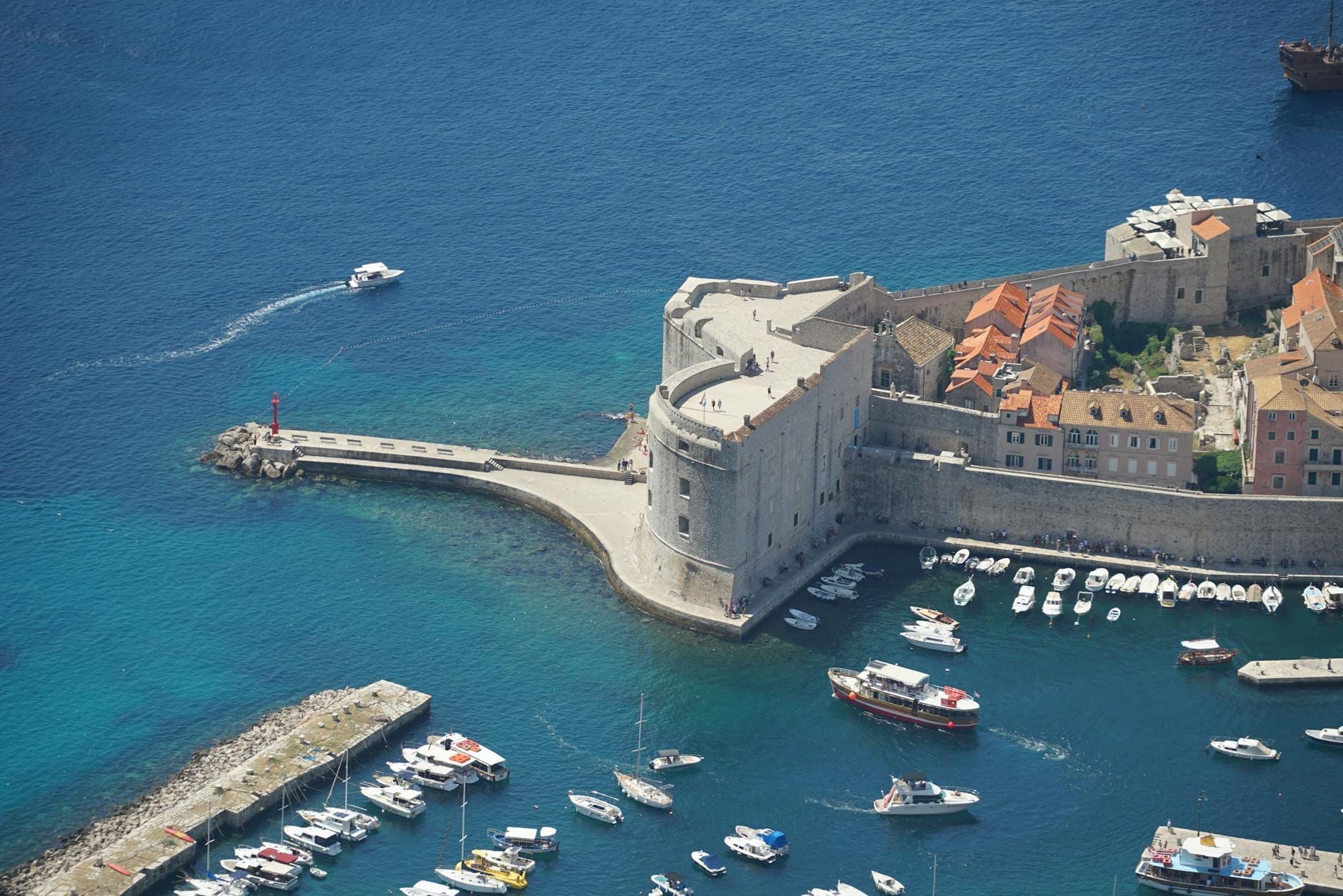 Vista aérea del Fuerte de San Juan en la entrada del Puerto Viejo de Dubrovnik, rodeado de aguas azules cristalinas y pequeñas embarcaciones, con las murallas de la ciudad y los tejados visibles al fondo.
