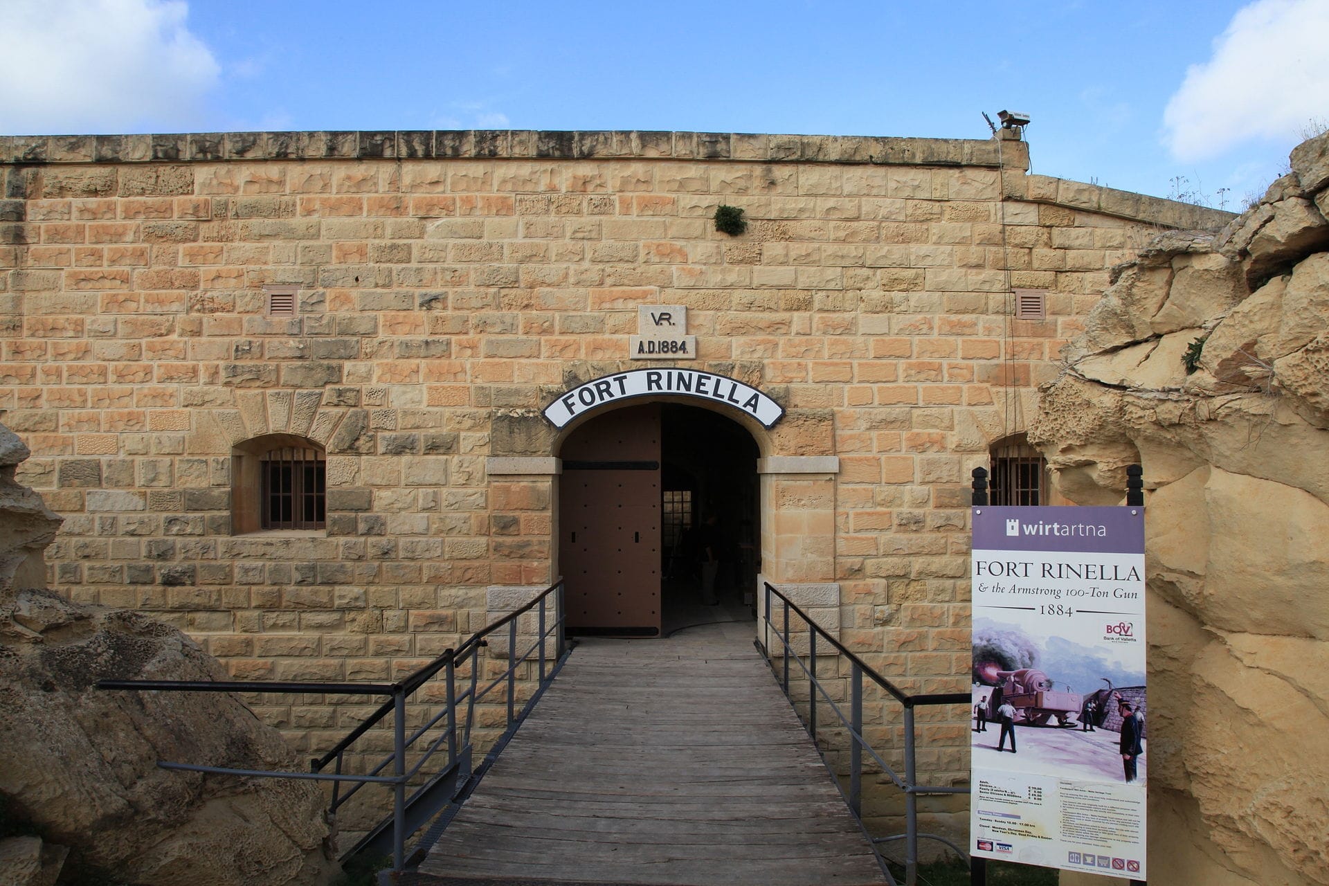 Ingresso di Fort Rinella con mura in pietra arenaria, portale ad arco e un cartello informativo sotto un cielo blu parzialmente nuvoloso a Kalkara, Malta.