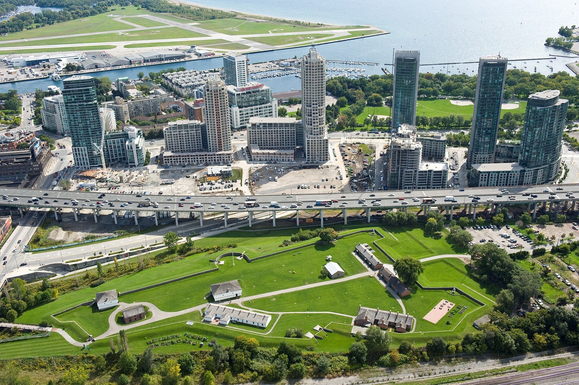 มุมมองทางอากาศของ Fort York National Historic Site พร้อมสนามหญ้าสีเขียว อาคารประวัติศาสตร์ และตึกระฟ้าใจกลางเมืองโตรอนโตในพื้นหลังในวันที่แดดจ้า