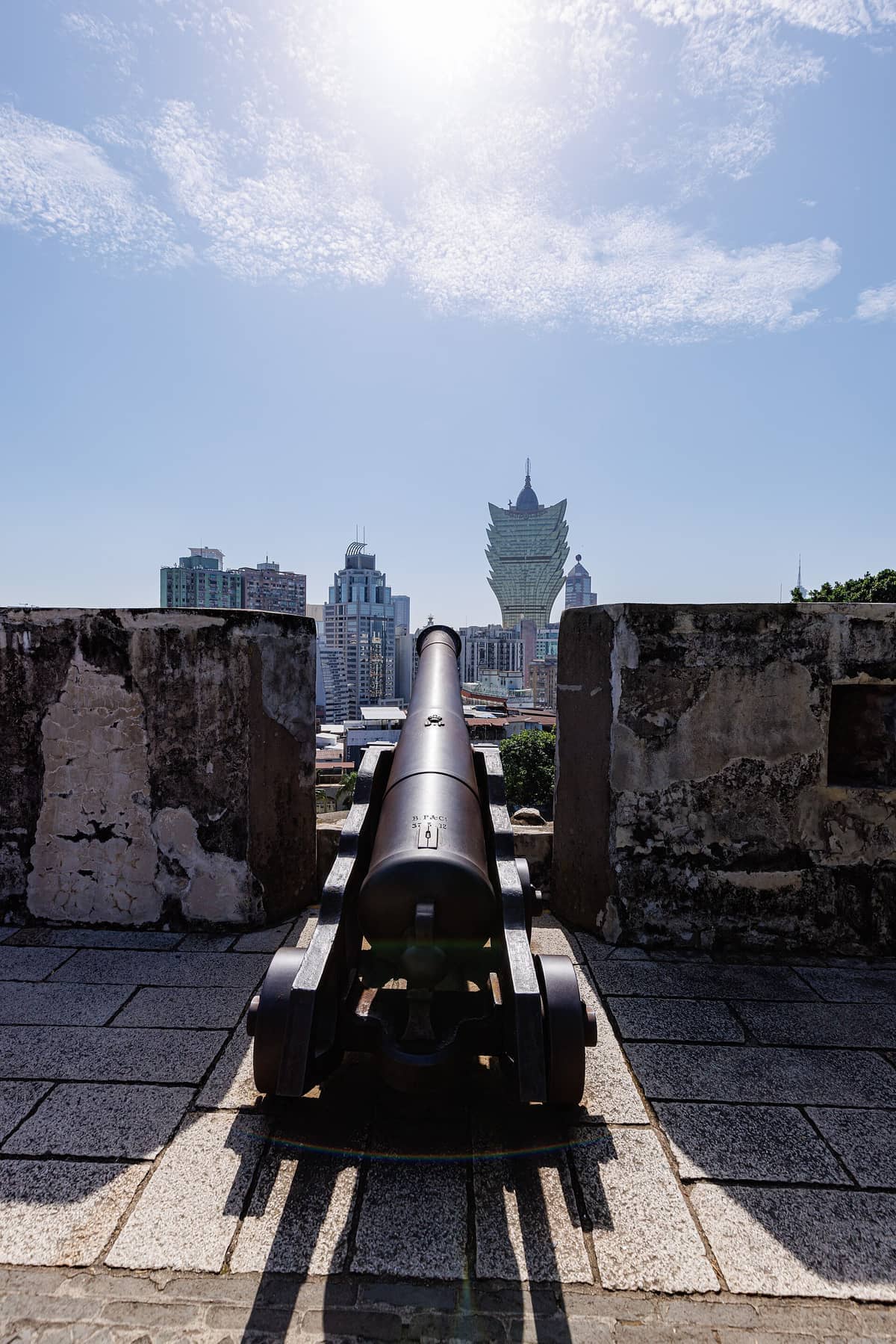 Historische Kanone auf der Fortaleza do Monte mit Blick auf die moderne Skyline von Macao unter hellem Mittagssonnenschein und blauem Himmel mit vereinzelten Wolken.