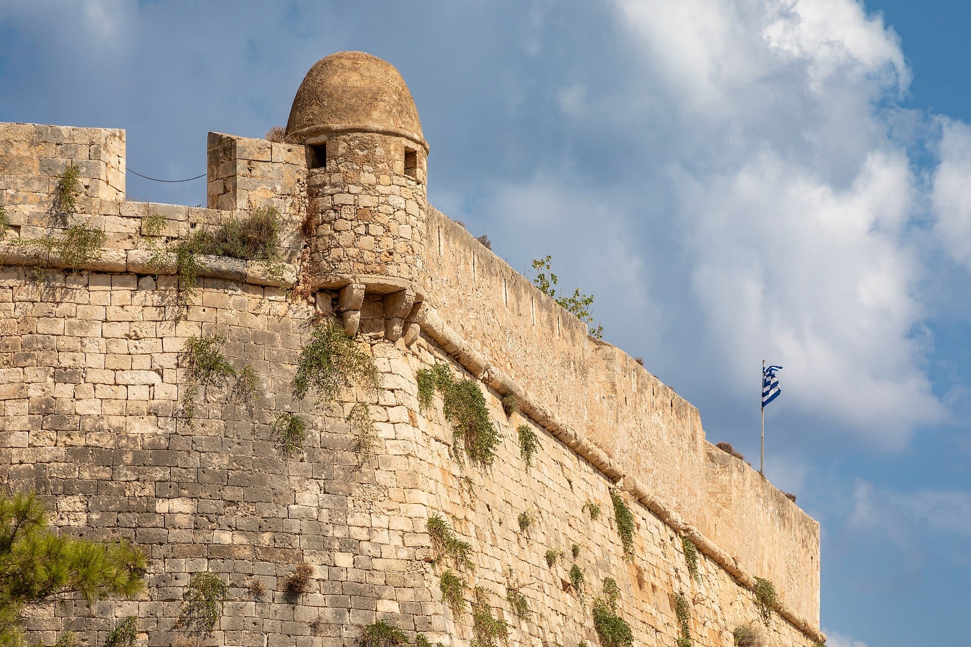 Mura in pietra e una torre di guardia della Fortezza di Rethymno con una bandiera greca su un cielo parzialmente nuvoloso.