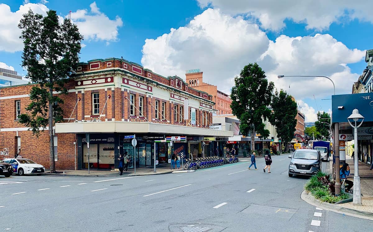 มุมมองถนนในย่าน Fortitude Valley, Brisbane เห็นตึกอิฐประวัติศาสตร์ ร้านค้า ต้นไม้ และผู้คนเดินเล่นบนฟุตปาธที่มีชีวิตชีวาภายใต้ท้องฟ้าสดใส