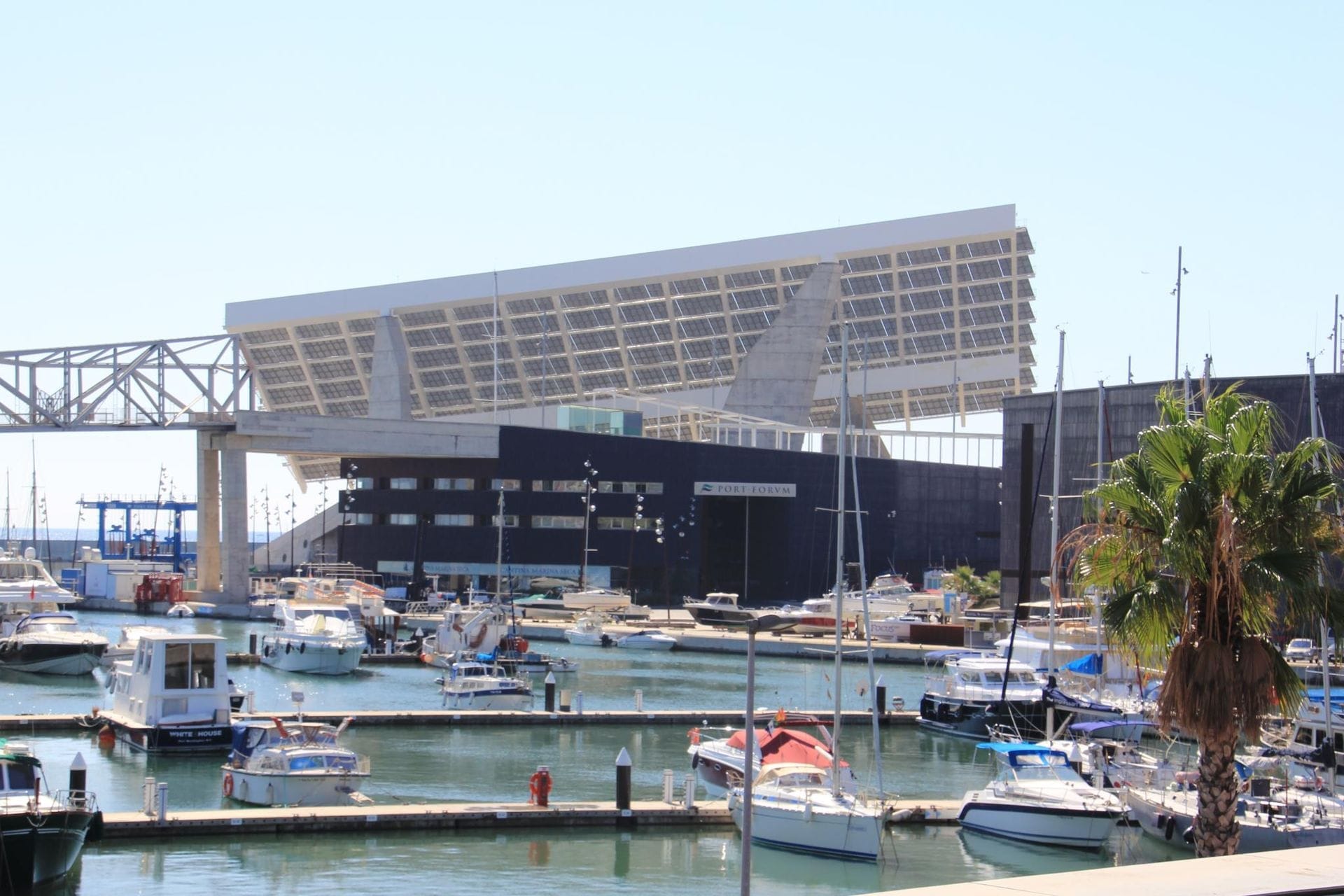 Modern solar panel structure at Port Fòrum Barcelona, with yachts docked in a calm marina and blue skies above.