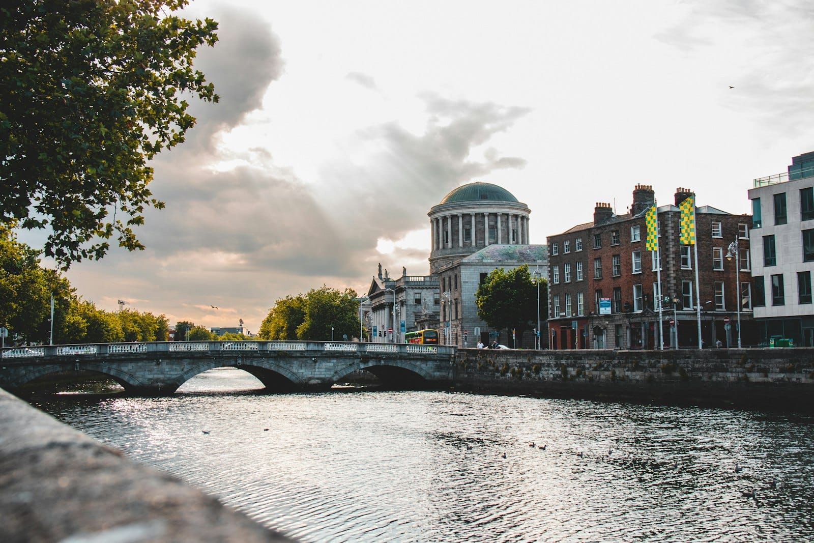 Veduta ampia del Four Courts di Dublino con la sua cupola e la facciata neoclassica sul Liffey, con un ponte in pietra sotto un cielo nuvoloso e drammatico.