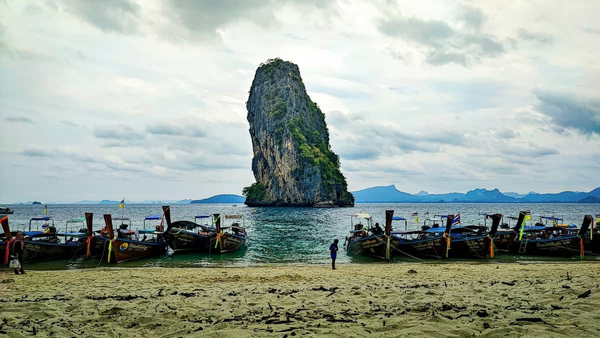 Botes de cola larga alineados en la playa de arena de Koh Poda, con el acantilado de piedra caliza de la isla emergiendo del agua turquesa bajo un cielo nublado.