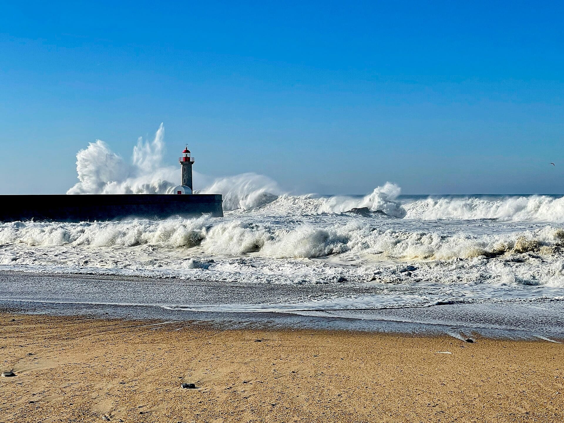Le onde si infrangono spettacolarmente contro il faro della spiaggia di Foz do Douro, con riva sabbiosa e cielo azzurro limpido a Porto, Portogallo.