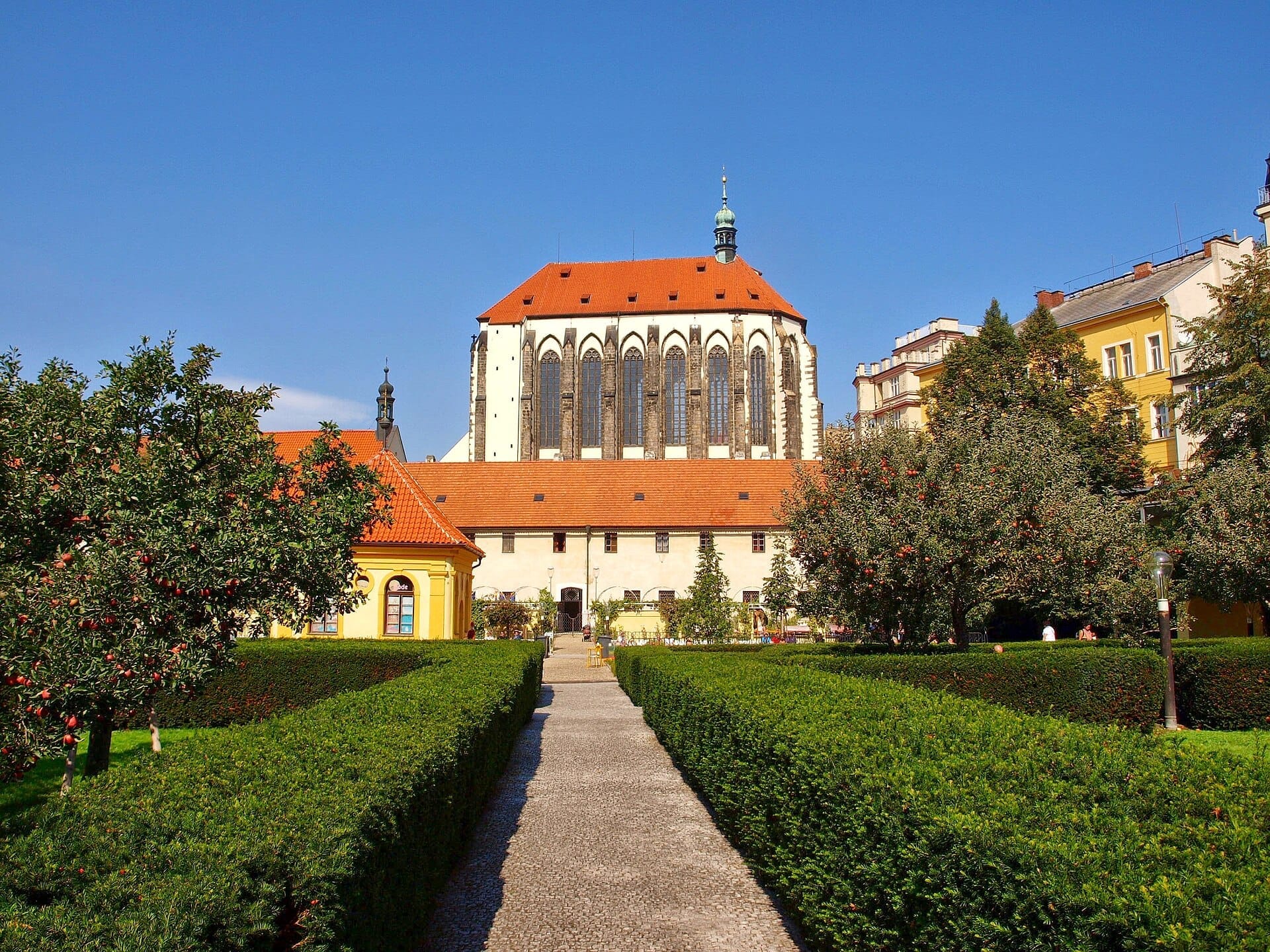 Franziskanergarten in Prag mit gepflegten Hecken, Obstbäumen und einer historischen Kirche unter einem klaren blauen Himmel.