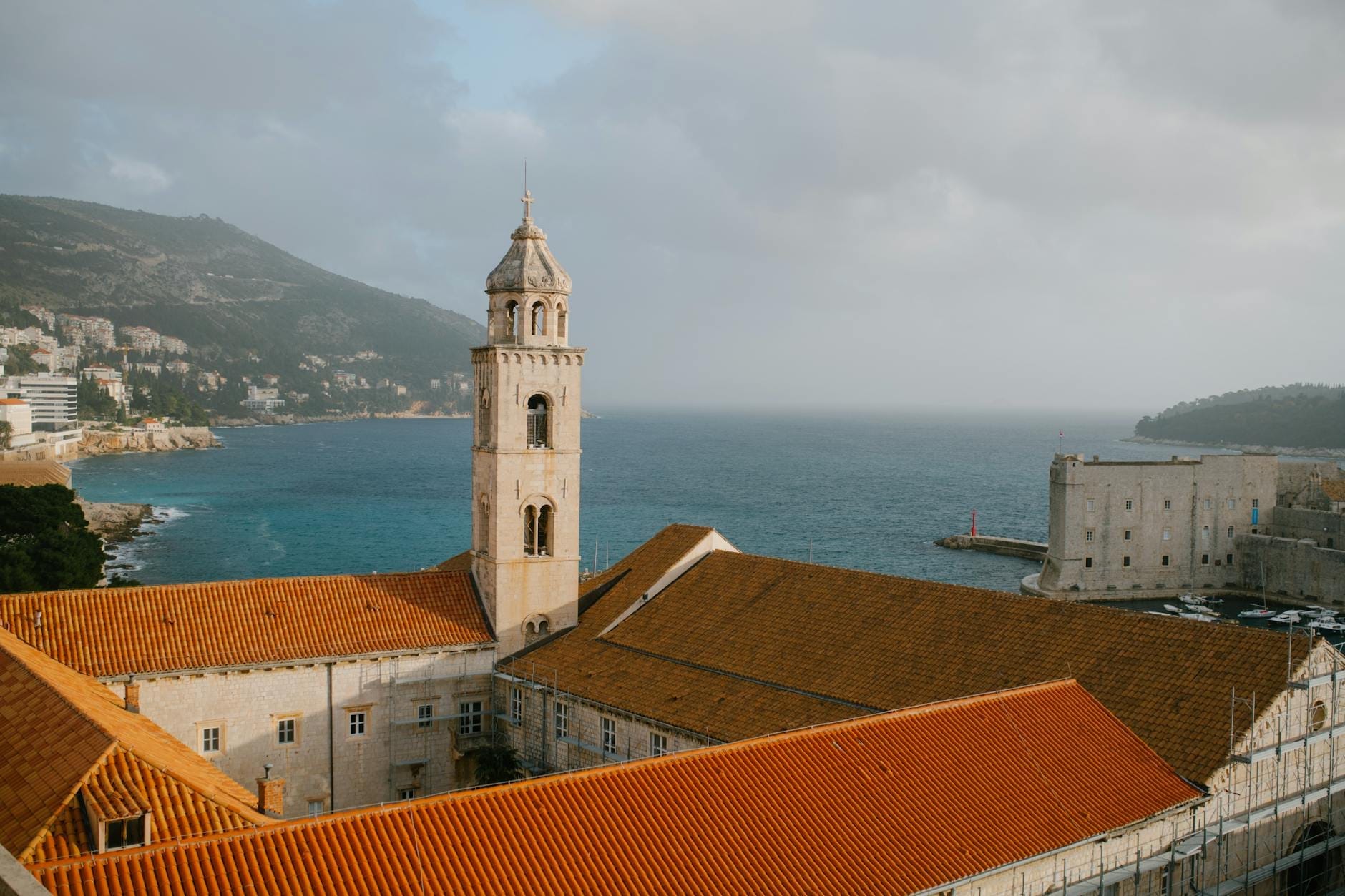 Vista panorámica del Monasterio Franciscano de Dubrovnik con su campanario y tejados de tejas naranjas sobre el mar Adriático, evocando el ambiente histórico de la Ciudad Vieja.