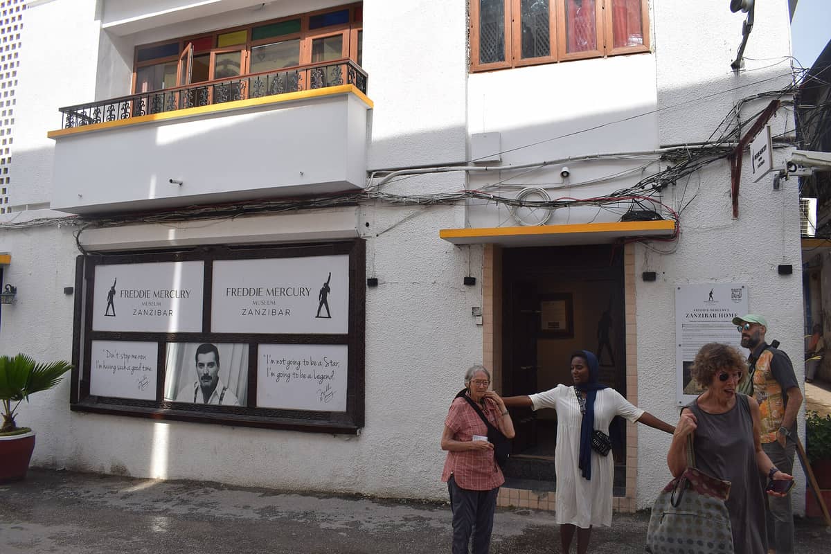 Esterno del Museo di Freddie Mercury a Stone Town, Zanzibar, con insegne che riportano il suo nome e alcune sue citazioni, e persone ferme davanti all'ingresso.