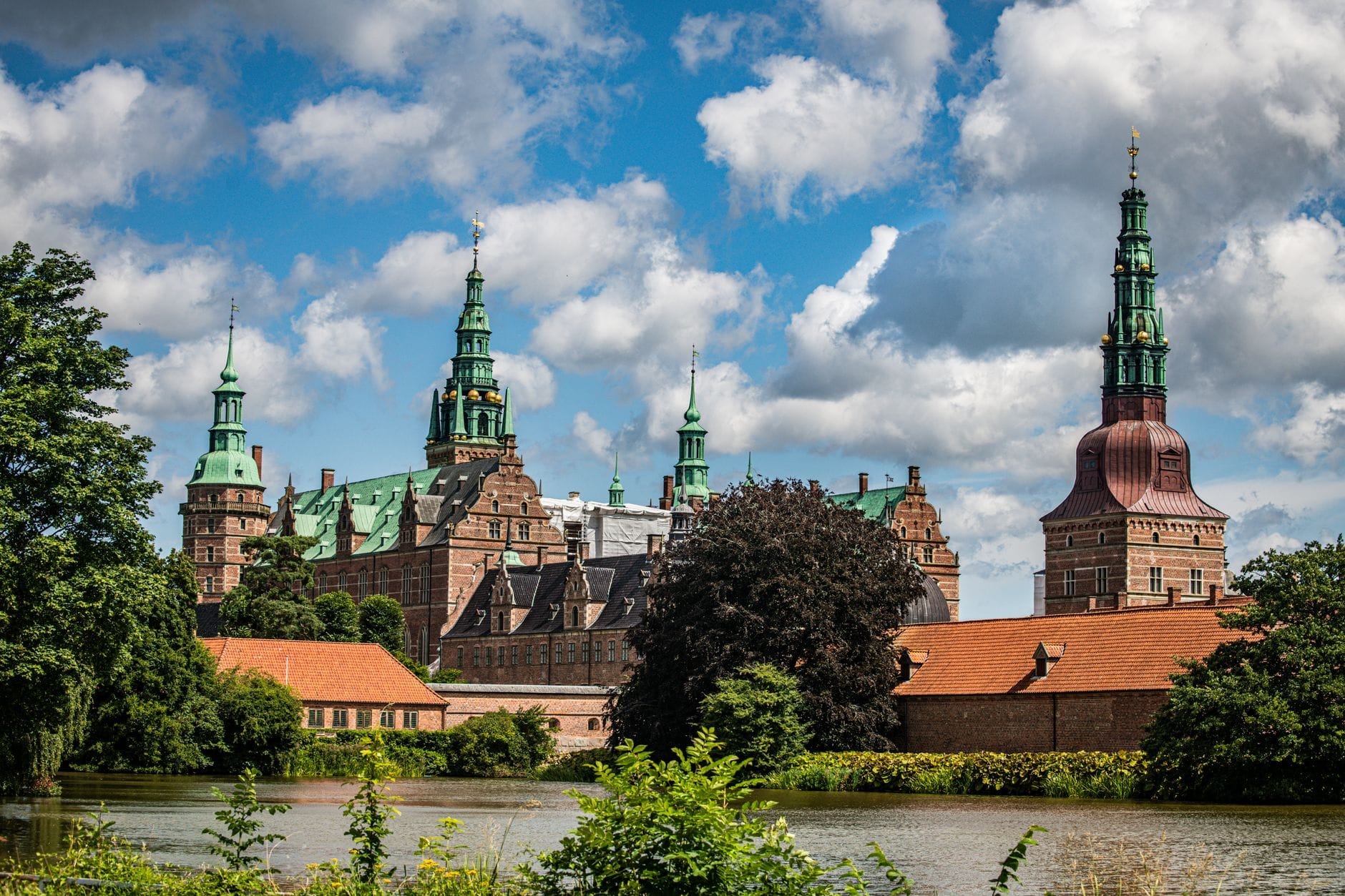 Il Castello di Frederiksborg in Danimarca visto dall'altra sponda del lago, con le torri in rame verde che si ergono sopra gli edifici in mattoni rossi sotto un cielo azzurro intenso con qualche nuvola.